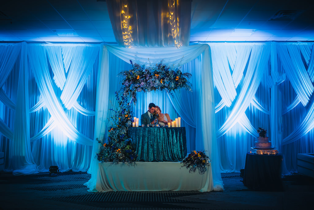 Florida Bride and Groom at Sweetheart Table Wedding Reception Portrait, Blue Linen Tablecloth, Blue Uplighting and Linen Drapery, Unique Creative Dramatic Blue, Purple, Orange Floral Arrangements | Tampa Bay Boutique Wedding Hotel Alba | Wedding Planner Special Moments Event Planning | Wedding Rentals Gabro Event Services | Styled Shoot