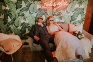 Fun Creative Bride and Groom with Sunglasses Lounging on Couch and Palm Tree Leaf Wallpaper Backdrop, Bride in Floral Lace and Illusion Halter Neckline Wedding Dress | Downtown St. Pete Wedding Venue Station House