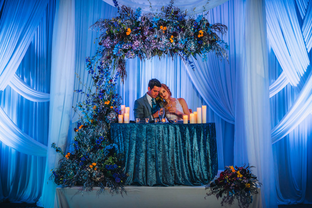 Florida Bride and Groom at Sweetheart Table Wedding Reception Portrait, Blue Linen Tablecloth, Blue Uplighting and Linen Drapery, Unique Creative Dramatic Blue, Purple, Orange Floral Arrangements | Tampa Bay Boutique Wedding Hotel Alba | Wedding Planner Special Moments Event Planning | Wedding Rentals Gabro Event Services | Styled Shoot
