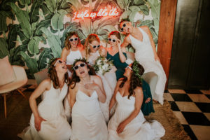 Fun Creative Bridal Party Portrait | Bridesmaids in Mix and Match White Dresses, Sunglasses, Maid of Honor in Emerald Green Dress with Palm Leaf Wallpaper Backdrop and Pink Neon Sign | Downtown St. Pete Wedding Venue Station House