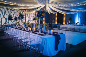 Dark Unique Modern Contemporary Dramatic Wedding Reception Decor, Long Feasting Tables with White Linens, String Lighting and White Linen Drapery, Tall Glass Vases with Unique Dark Blue, Purple Floral Centerpieces, Creative Artsy Wall Projection, Ghost Acrylic Clear Chiavari Chairs, Blue Table Runner | Tampa Bay Boutique Hotel Wedding Venue Hotel Alba | Wedding Planner Special Moments Event Planning | Wedding Rentals and Chair Decor Gabro Event Services | Styled Shoot