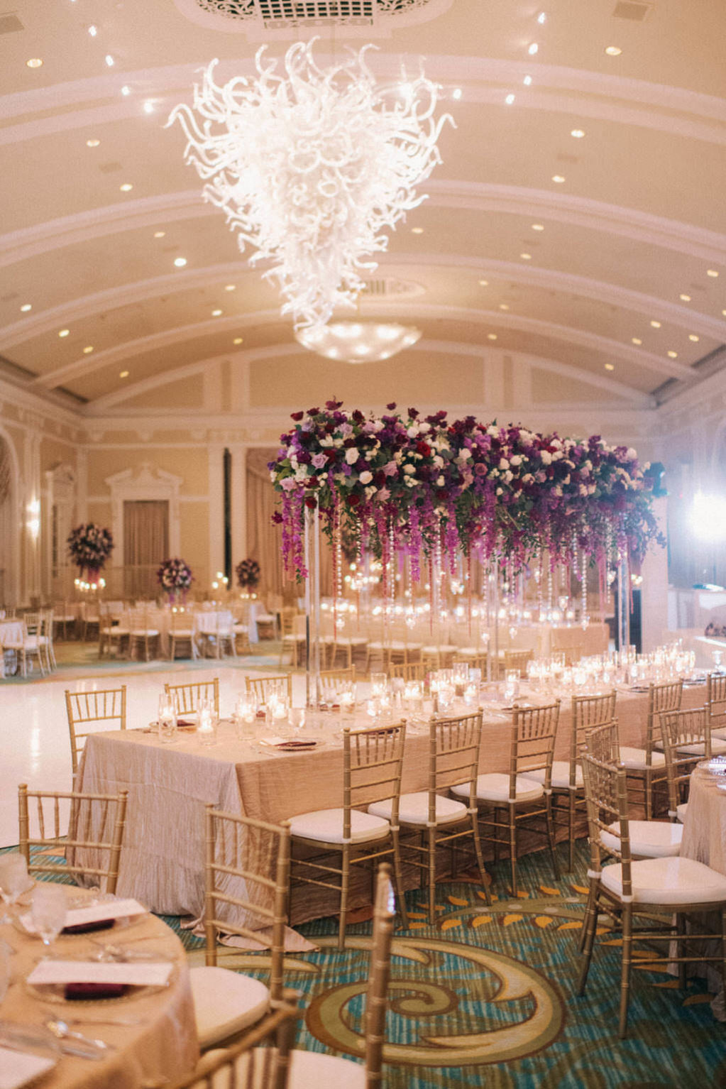 Elegant, Extravagant Wedding Reception Decor Portrait, Long Table with Gold Linen, Gold Chiavari Chairs, Tall Purple, Red, Plum, Lilac, Greenery and Purple Hanging Amaranthus Floral Centerpiece, Artistic Glass Chandelier | Downtown St. Pete Hotel Ballroom Wedding Venue The Vinoy Renaissance