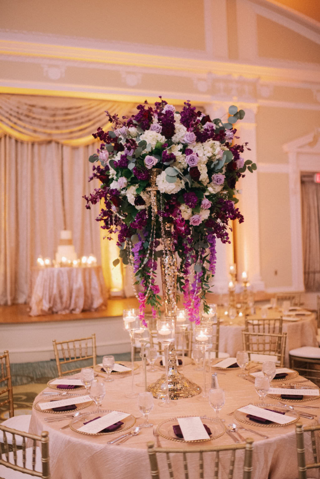 Elegant and Luxurious Wedding Reception Decor Portrait, Round Table with Gold Linen, Tall Purple, Plum, Lilac, Ivory and Hanging Amaranthus and Draped Crystals Floral Centerpiece