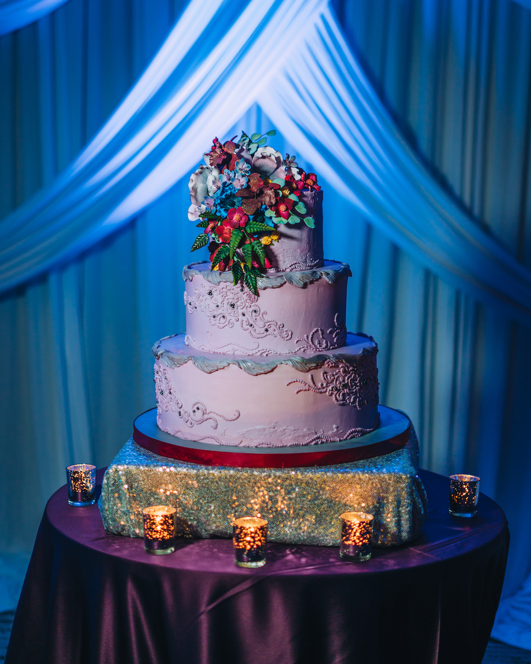 Unique Three Tier Purple Wedding Cake with Floral Icing Accents, Sugar Flower Red, Blue, Purple, Yellow and Greenery Cake Topper, Gold Glitter Cake Stand and Round Table with Purple Tablecloth, Blue Uplighting and Linen Drapery | Tampa Wedding Baker Alessi Bakery | Wedding Planner Special Moments Event Planner | Styled Shoot