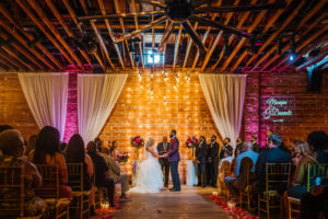 Florida Bride and Groom During Vow Exchange, Romantic Indoor Wedding Ceremony Decor, Exposed Red Brick Wall Ceremony Backdrop with Vintage Hanging Lights, White Draping, Gold Chiavari Chairs, Blush Pink Roses, Burgundy Flowers, Plum Hibiscuses, Quartz and Magenta Florals, Dark Greenery and Gold Centerpiece | Historic Tampa Bay Destination Industrial Wedding Venue NOVA 535, American Basketball Player Davante Gardner