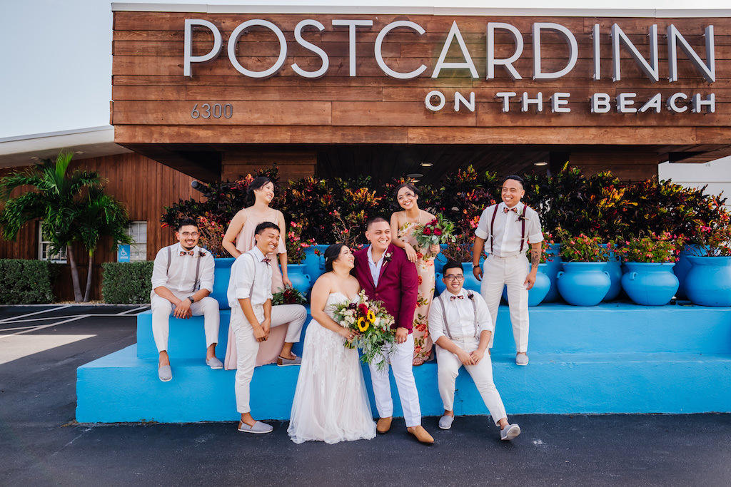Fun Bride Holding Colorful Organic Yellow Sunflowers, Greenery, and Red Floral Bridal Bouquet, Groom in Burgundy Red Suite Jacket and White Dress Pants, Groomsmen in White Capri Pants, Suspenders and Bowties, Bridesmaid in Floral Floor Length Dress, Wedding Party Portrait Outside St. Petersburg Wedding Venue Postcard Inn on the Beach