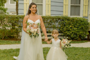 Garden Boho St. Pete Bride in Lace V Neckline with Spaghetti Straps Stella York Wedding Dress Holding King Protea, Ivory and Blush Pink Roses, Eucalyptus and Greenery Floral Bouquet with Daughter Flower Girl in White and Blush Tulle Skirt Holding Garden Ivory, Pink Roses and Greenery Floral Bouquet Walking Down Wedding Ceremony Portrait | Tampa Bay Wedding Hair and Makeup LDM Beauty Group