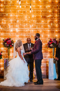 Florida Bride and Groom During Vow Exchange, Romantic Garden Indoor Wedding Ceremony Decor, Exposed Red Brick Wall Ceremony Backdrop with Vintage Hanging Lights, Blush Pink Roses, Burgundy Flowers, Plum Hibiscuses, Quartz and Magenta Florals, Dark Greenery and Gold Centerpiece | Historic Tampa Bay Destination Industrial Wedding Venue NOVA 535, American Basketball Player Davante Gardner