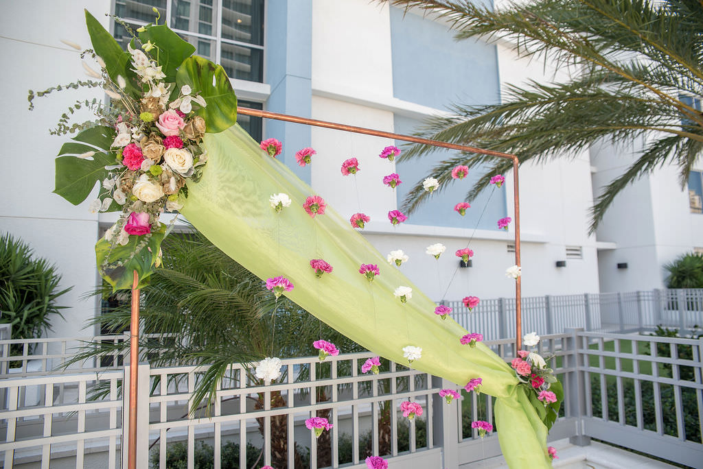 Colorful Tropical Wedding Ceremony Decor, Copper Arch with Lime Green Draping, Fuschia Pink and White Hanging Carnations, Ivory, Pink and Metallic Gold Painted Roses with Monstera Palm Leaves Floral Arrangement | Tampa Bay Wedding Photographer Kristen Marie Photography | Hotel Wedding Venue Hyatt Place Downtown St. Pete | Wedding Florist Brides N Blooms | Outside the Box Event Rentals