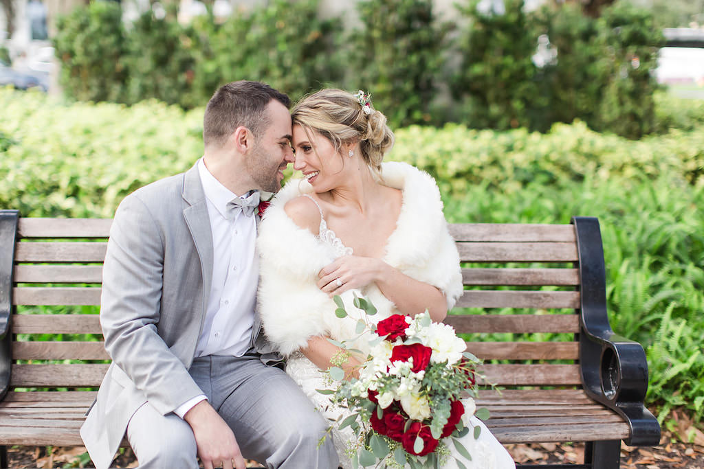Christmas Inspired Bride and Groom Sitting on Bench, Bride Wearing Winter Wedding Style White Fur Coat, Holding Rustic White Floral Bouquet with Red Roses, and Greenery, in Downtown St. Pete Straub Park | Tampa Bay Wedding Photographers Shauna and Jordon Photography