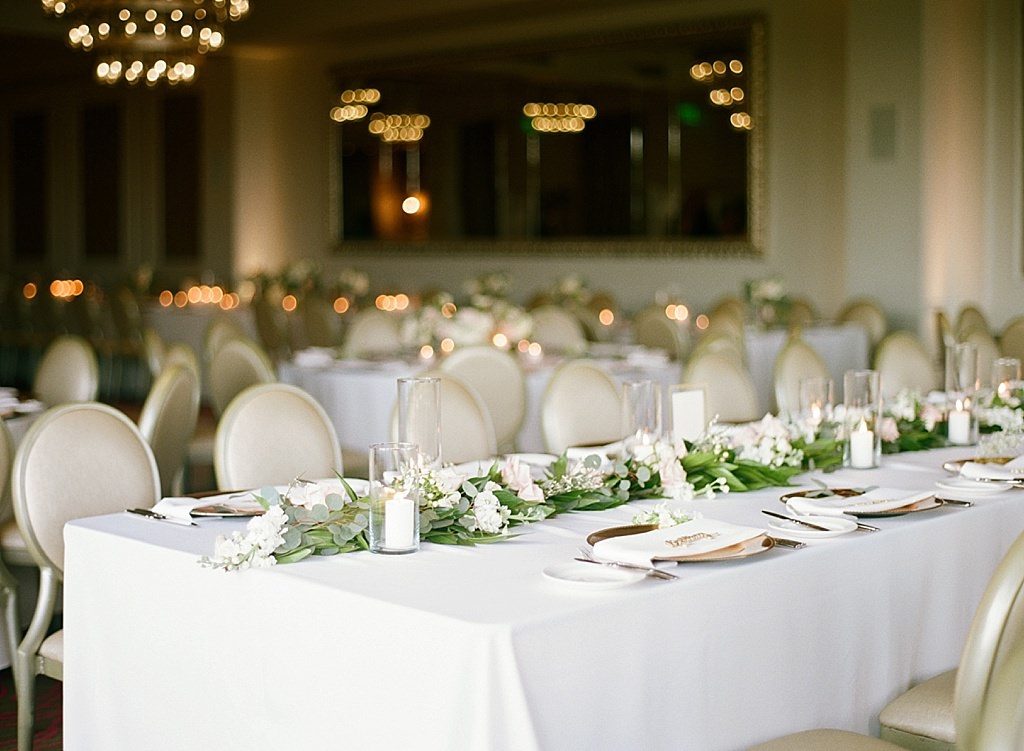Formal Modern Wedding Reception Decor, Long Feasting Table with White Linen, Eucalyptus Greenery Garland, White Rose Florals, Gold Chargers and Glass Hurricane Candles | Downtown St. Pete Ballroom Wedding Venue The Birchwood