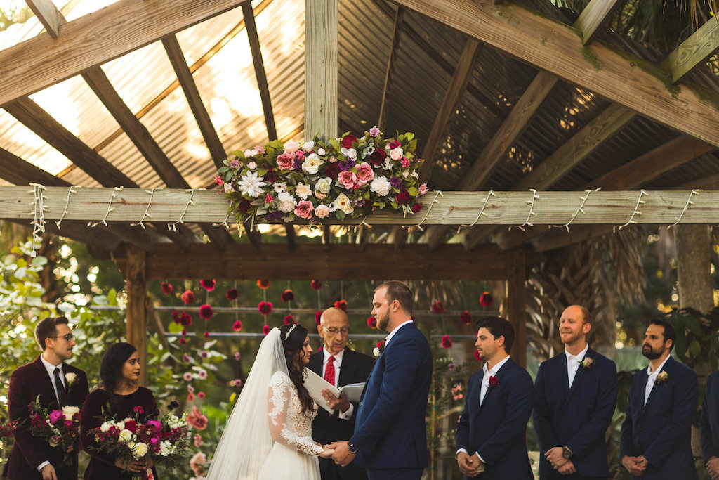 Romantic Classy Boho Chic Wedding Ceremony Bride and Groom Exchanging Vows Portrait Under Gazebo with White, Pink and Purple Roses with Greenery, Hanging Red Flowers