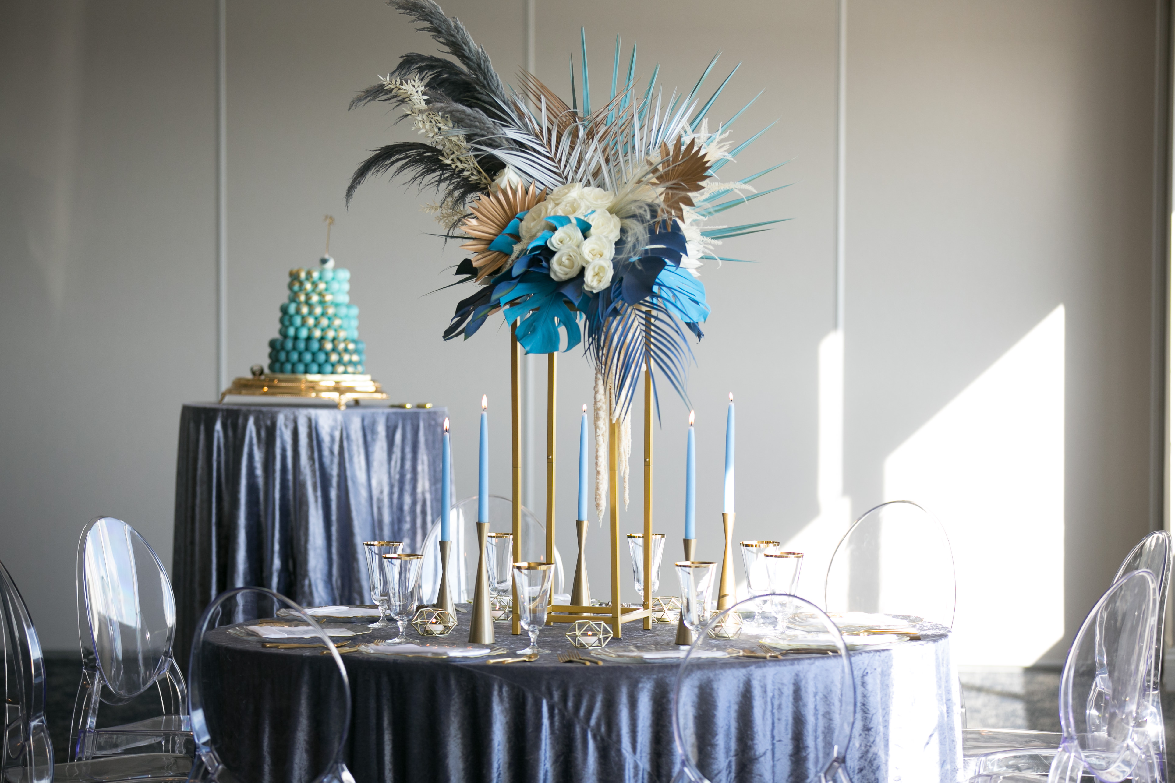 Whimsical Unique Tropical Wedding Reception Decor, Round Tables with Midnight Blue Linen, Tall Gold Candlesticks with Blue Candles, Tall Gold Geometric Stands with White Roses, Blue Monstera and Palm Tree Leaves and Feathers and Ghost Chair Seating | Tampa Wedding Chair Rentals Gabro Event Services | Velvet Navy Blue Table Linens Kate Ryan Event Rentals | Wedding Photographer Carrie Wildes Photography | Wedding Planner and Florist John Campbell Weddings | Tampa Wedding Venue Centre Club