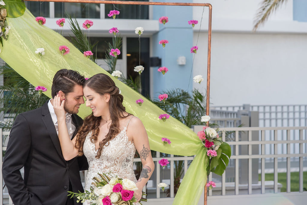 Modern Bride in Low V Neckline Lace and Illusion with Spaghetti Strap Wedding Dress and Groom in Black Tuxedo Wedding Portrait, Colorful Tropical Wedding Ceremony Decor, Copper Arch, Fuschia Pink and White Hanging Carnations, Lime Green Draping, Pink and Ivory Roses, Monstera Palm Leaf Floral Arrangement | Tampa Bay Wedding Photographer Kristen Marie Photography | Hotel Wedding Venue Hyatt Place Downtown St. Pete | Wedding Florist Brides N Blooms | Wedding Attire Nikki's Glitz and Glam Boutique | Outside the Box Event Rentals | Wedding Hair and Makeup LDM Beauty Group