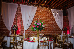Romantic, Modern Wedding Decor and Reception, Tall Gold Centerpieces with Lush Floral and Greenery, Blush Pink Roses, Burgundy Flowers, Plum Hibiscuses, Quartz and Magenta Blooms, Chiavari Chairs and Chargers, Round Tables with White Linen and Draping, Exposed Red Brick Wall with Vintage Hanging Indoor Lighting | Unique Florida Industrial Wedding Venue NOVA 535 in Downtown St. Pete | Tampa Event Caterer Olympia Catering
