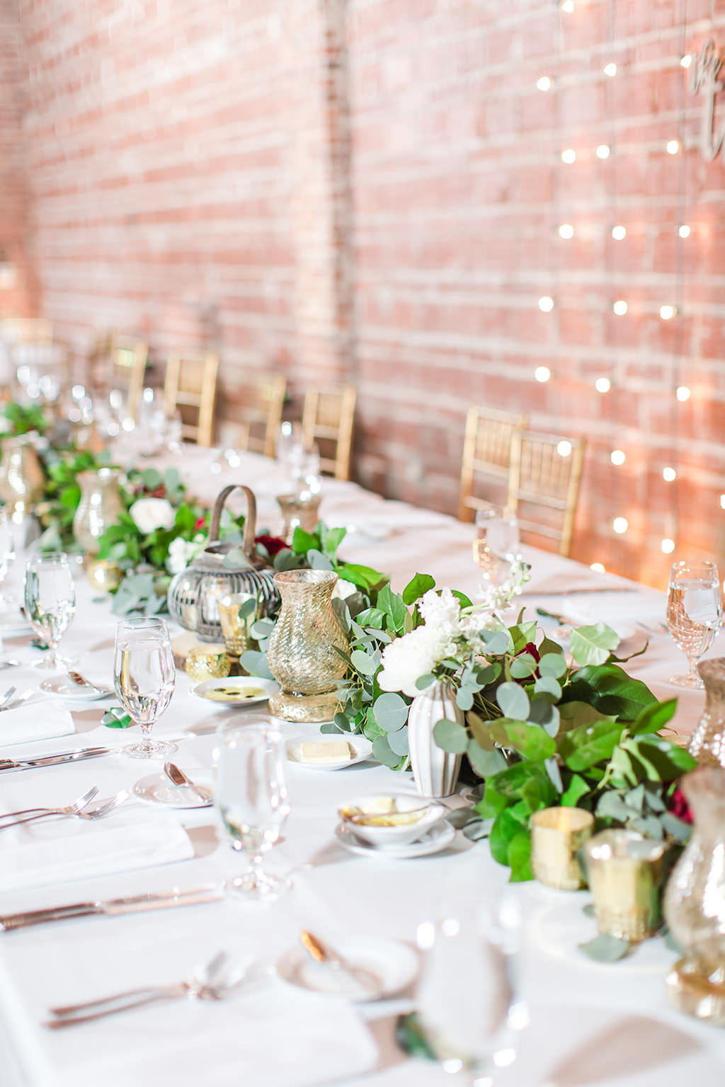 Rustic Chic Inspired Wedding Decor at Feasting Table, Romantic Greenery Eucalyptus Leaves with White and Red Floral Garland Centerpiece, Nature Inspired Lanterns, Mercury Glass Vases, Gold Chiavari Chairs, Against Exposed Red Brick Wall with Indoor String Lighting | Tampa Bay Unique Wedding Venue NOVA 535 | St. Pete Luxury Wedding Photographers Shauna and Jordon Photography