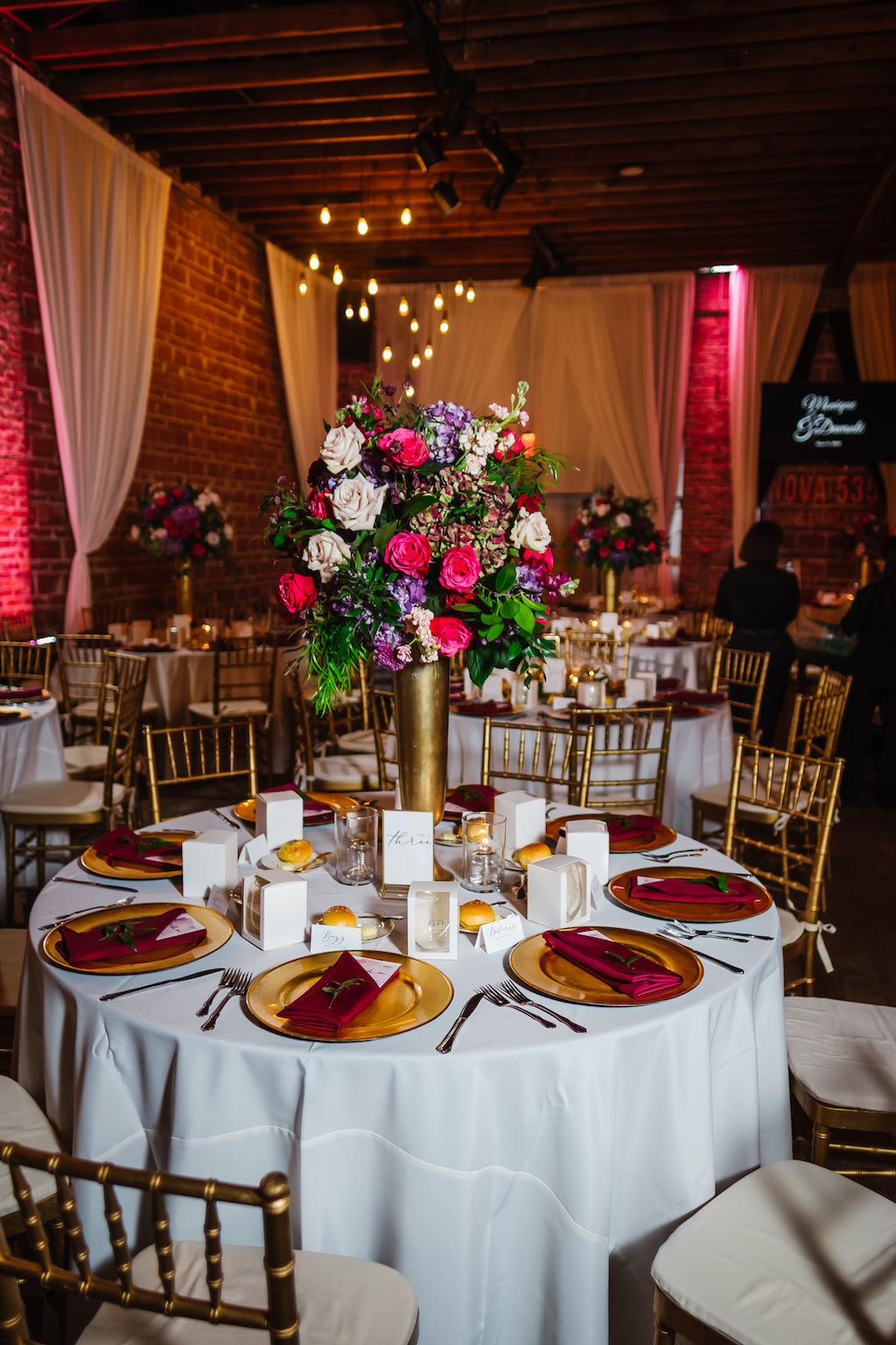 Romantic, Garden Modern Wedding Decor and Reception, Tall Gold Centerpieces with Lush Floral and Greenery, Blush Pink Roses, Burgundy Flowers, Plum Hibiscuses, Quartz and Magenta Blooms, Chiavari Chairs and Chargers, Round Tables with White Linen and Draping, Exposed Red Brick Wall with Vintage Hanging Indoor Lighting | Unique Florida Industrial Wedding Venue NOVA 535 in Downtown St. Pete