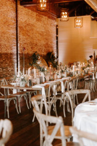 Boho Whimsical Wedding Reception Decor, Long Wooden Feasting Table with Floral Arrangements and Hurricane Glass Candle Holders, Wooden Crossback Chairs | Tampa Bay Wedding Planner Coastal Coordinating | Industrial Wedding Venue Armature Works