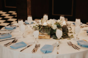 Elegant Wedding Reception Decor, Round Table with White Tablecloth, Dusty Blue Linen Napkins, Acrylic Clear and Gold Table Number, White Roses and Eucalyptus Floral Centerpiece, Hurricane Glass Cylinder with Floating Candles