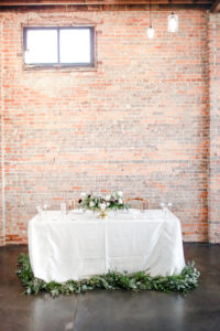 Industrial Boho Chic Wedding Reception Decor, Sweetheart Table with White Linen, Greenery Garland, White Rose and Dark Purple, Plum Roses in Gold Vase Low Centerpiece and Red Brick Backdrop | Tampa Wedding Photographer Lifelong Photography Studio | Wedding Planner Special Moments Event Planning | Wedding Tables, Chairs and Linen Rentals Gabro Event Services | Unique Wedding Venue CAVU
