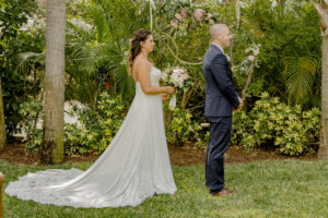 Florida Bride and Groom First Look Wedding Portrait, Bride in Lace Train A-Line Stella York Wedding Dress Holding Garden Inspired Bridal Floral Bouquet, Two Gold Hanging Hoops with King Proteas and Greenery Floral Arrangements | Tampa Bay Wedding Hair and Makeup LDM Beauty Group