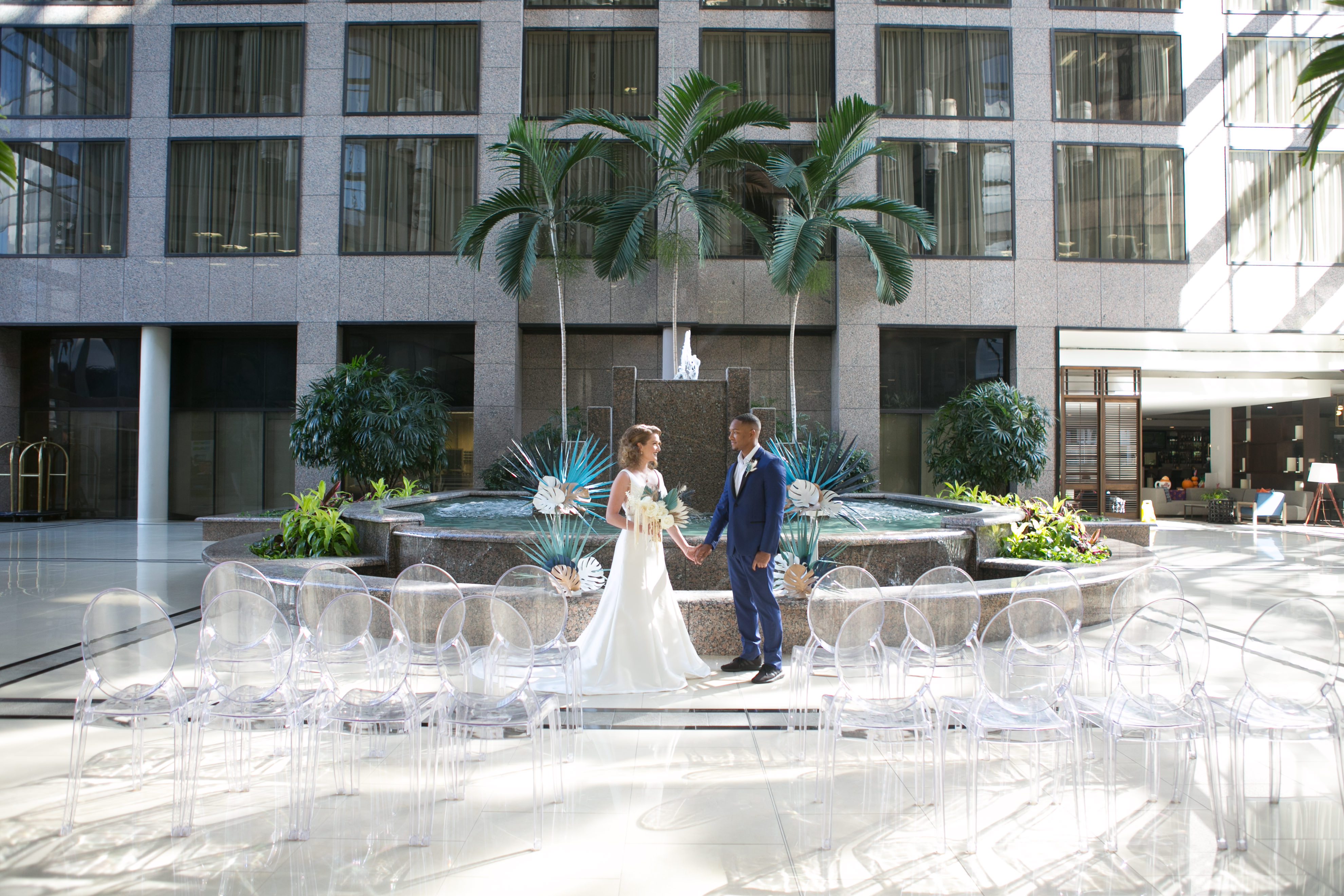 Unique Tropical Wedding Ceremony Decor, Acrylic Ghost Chair Seating, Bride and Groom Standing in Front of Fountain with Palm Tree Leave Arrangements | Tampa Wedding Chair Rentals Gabro Event Services | Wedding Planner and Florist John Campbell Weddings | Wedding Photographer Carrie Wildes Photography | Tampa Wedding Venue Centre Club