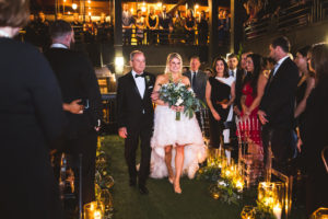 Modern Bride in High Low Ruffled Skirt Ines Di Santo Wedding Dress Holding Greenery and White Floral Bouquet with Father Walking Down the Wedding Ceremony Aisle, Romantic Candles, New Years Eve Nighttime Rooftop Wedding | St. Petersburg Wedding Venue Station House