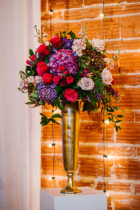 Romantic Garden Indoor Wedding Decor, Exposed Red Brick Wall Ceremony Backdrop with Vintage Hanging Lights, White Draping, Gold Chiavari Chairs, Blush Pink Roses, Purple Hydrangeas, Burgundy Flowers, Plum Hibiscuses, Quartz and Magenta Florals, Dark Greenery and Gold Arrangement | Historic Downtown St. Pete Industrial Wedding Venue NOVA 535