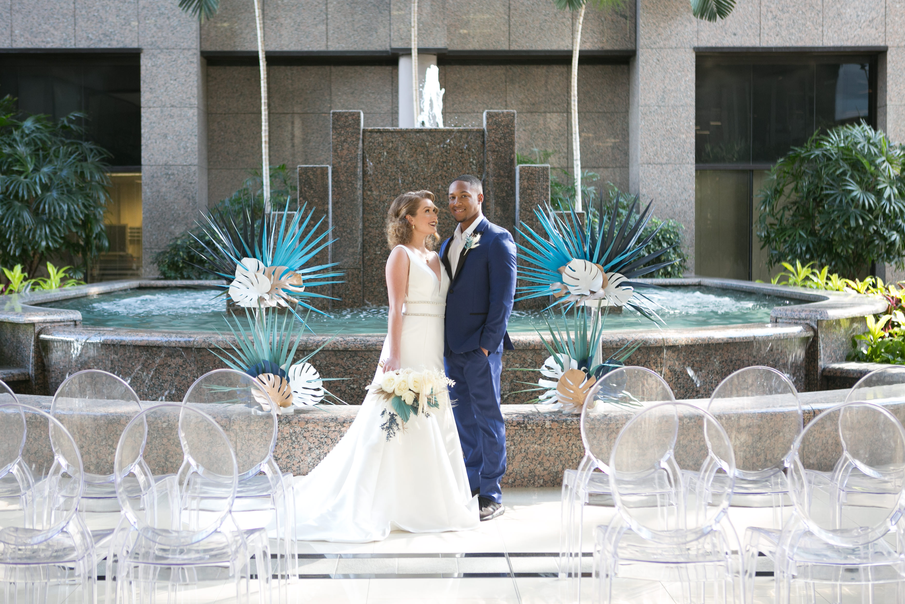 Bride and Groom Wedding Ceremony Portrait, Bride in Classic A-Line V Neck Wedding Dress Holding Ivory Roses and Greenery Bridal Floral Bouquet, Groom in Blue Suit, Green, Ivory and Gold Palm Tree and Monstera Leaves and Ghost Chair Wedding Ceremony Seating Fountain Portrait | Tampa Wedding Chair Rentals Gabro Event Services | Wedding Photographer Carrie Wildes Photography | Wedding Venue Centre Club | Wedding Planner and Florist John Campbell Weddings | Wedding Attire Truly Forever Bridal