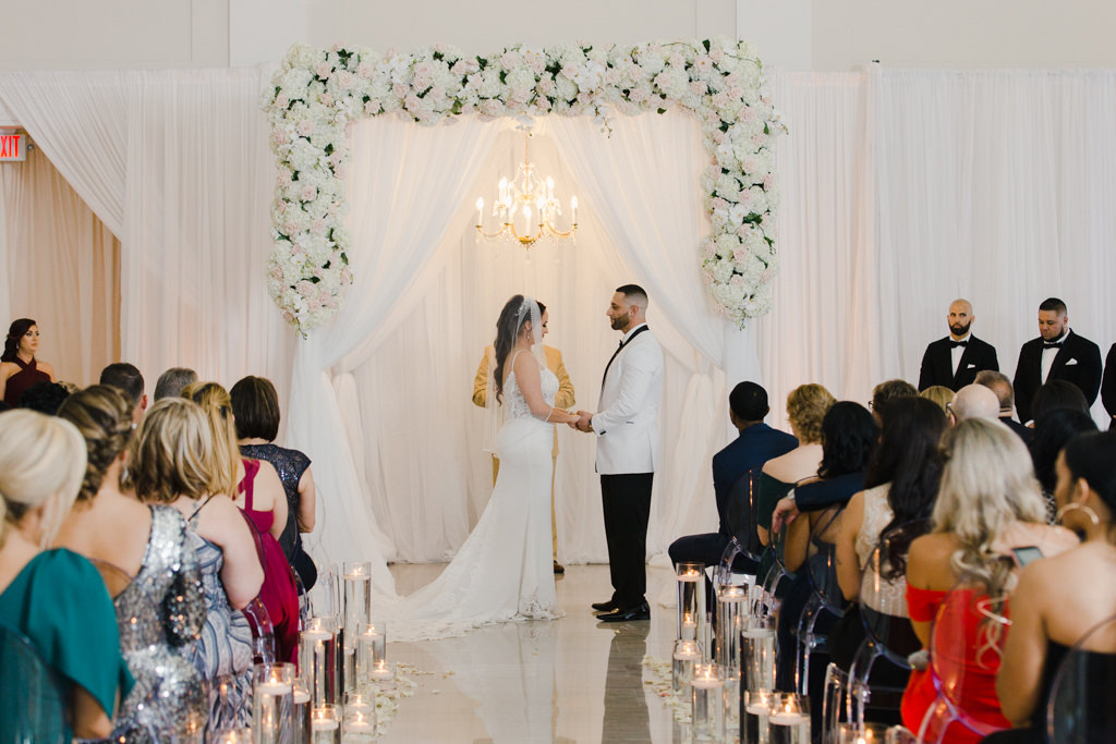Modern Romantic Bride and Groom Exchanging Vows During Wedding Ceremony, White Draping Backdrop with White and Blush Pink Floral Arrangement Arch and Gold Crystal Hanging Chandelier | Tampa Bay Wedding Venue The Vault | Wedding Florist and Rentals Gabro Event Services