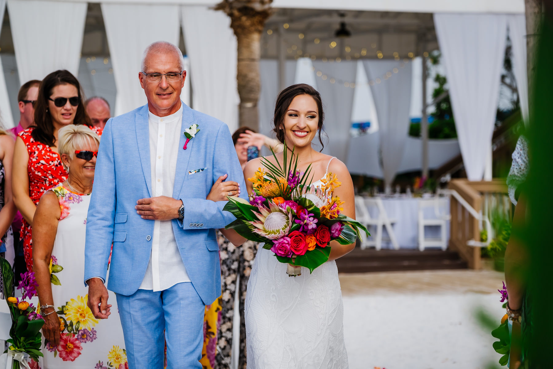 Tropical Clearwater Beach Bride Walking Down the Wedding Ceremony Aisle with Father Holding King Protea, Purple, Red and Orange Roses, Purple Orchids, Palm Fronds and Monstera Leaf Floral Bridal Bouquet