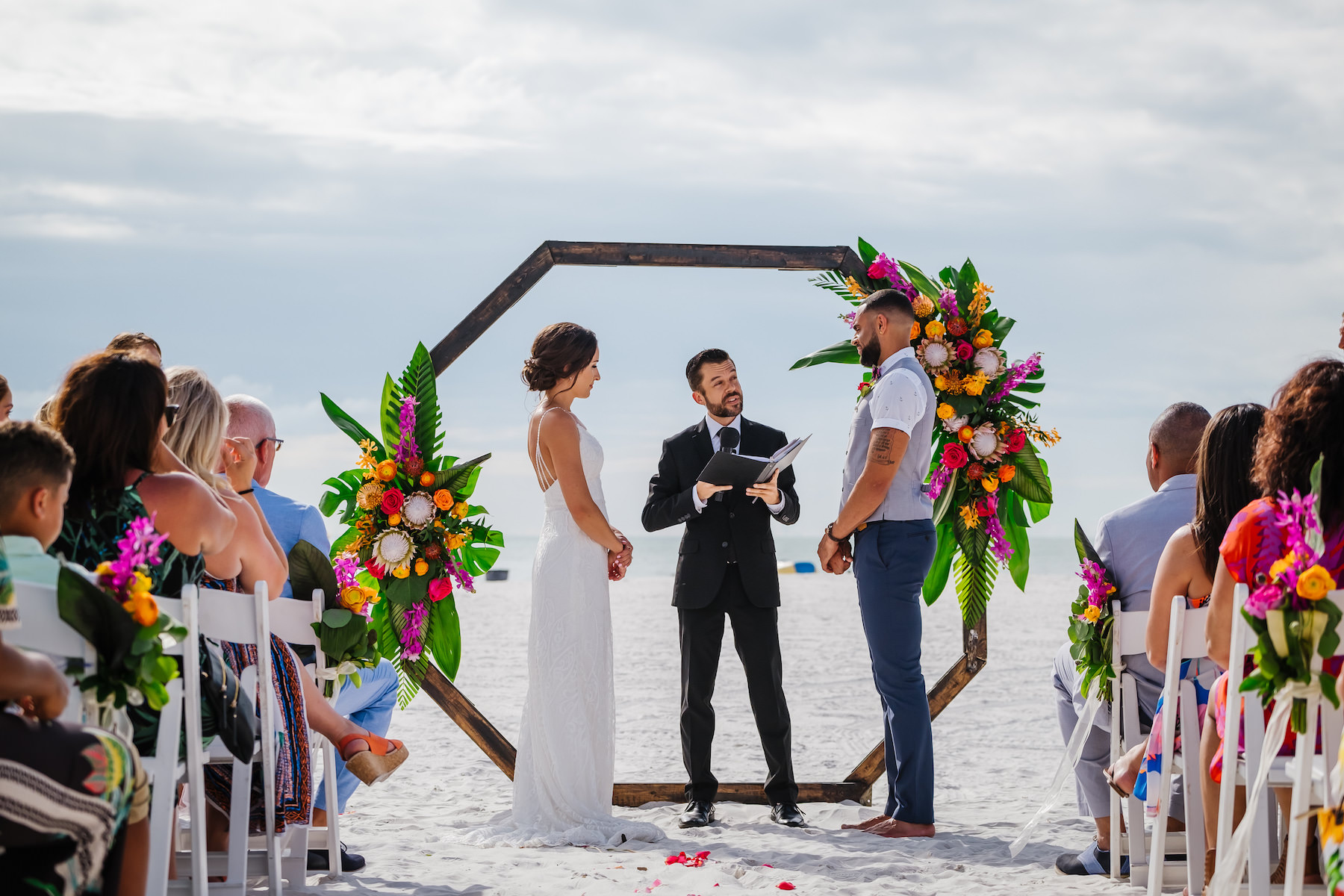 Clearwater Beach Bride and Groom Exchanging Wedding Ceremony Vows Waterfront, Geometric Octagonal Wooden Arch with Tropical King Proteas, Orange, Pink and Red Roses, Purple Orchids, Palm Fronds and Monstera Palm Leaves Floral Arrangements on Beach Portrait | Tampa Bay Waterfront Beach Hotel Wedding Venue Hilton Clearwater Beach