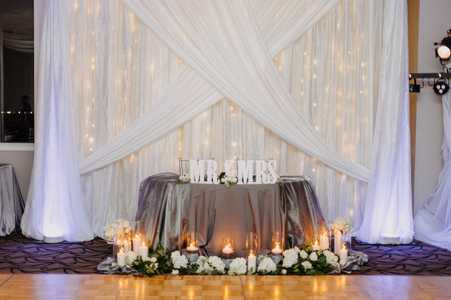 Classic Elegant Wedding Reception Decor, White Draping Backdrop with String Lights, Silver Linen on Sweetheart Table with Mr and Mrs Sign, White Hydrangeas, Glass Cylinder Candles | Tampa Bay Wedding Venue Centre Club
