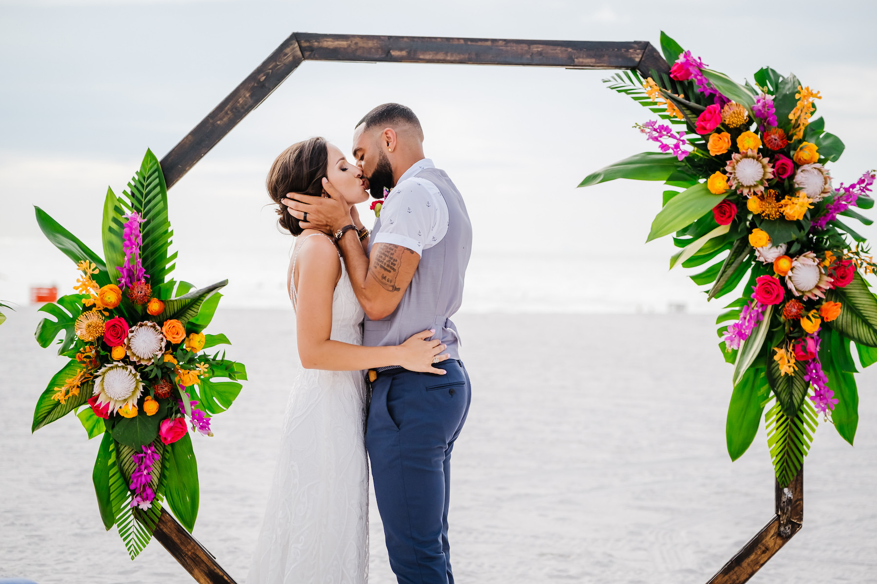 Clearwater Beach Intimate Kissing Bride and Groom Portrait, Geometric Octagonal Wooden Arch with Tropical King Proteas, Orange, Pink and Red Roses, Yellow and Purple Accents, Palm Fronds and Monstera Leaves Floral Arramngements, Beachfront and Waterfront Wedding