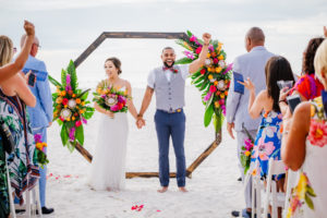 Clearwater Beach Bride and Groom Excitement Wedding Portrait After Saying I Do's, Geometric Octagonal Wooden Arch with Tropical Palm Fronds, Monstera Leaves, Pink, Orange and Red Roses, Yellow and Purple Floral Arrangements | Tampa Bay Waterfront Beach Hotel Wedding Venue Hilton Clearwater Beach