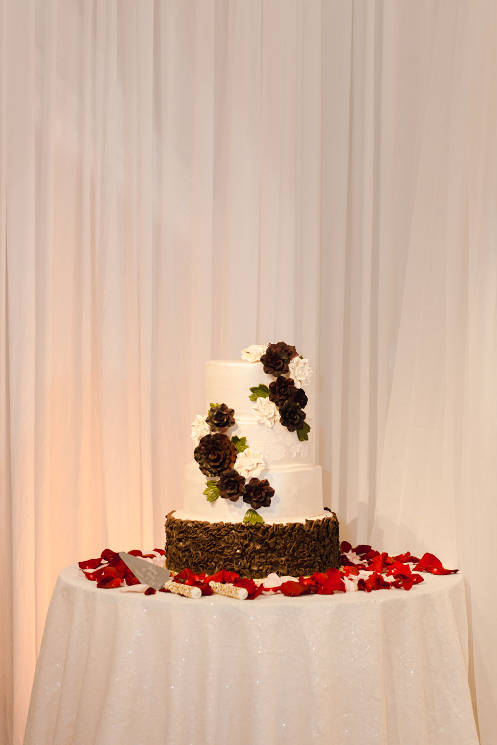 Three Tier White and Chocolate Brown Ruffle Wedding Cake with Cascading Sugar White and Brown Roses on Round Table with White Sparkle Linen and Red Rose Petals and White Drapery Backdrop