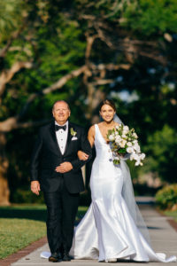 Bride in Classic Silhouette V Neckline Wedding Dress Holding White Orchids and Greenery Floral Bouquet Walking Down the Wedding Ceremony Aisle with Father | Tampa Bay Wedding Planner NK Productions | Wedding Hair and Makeup Femme Akoi Beauty Studio