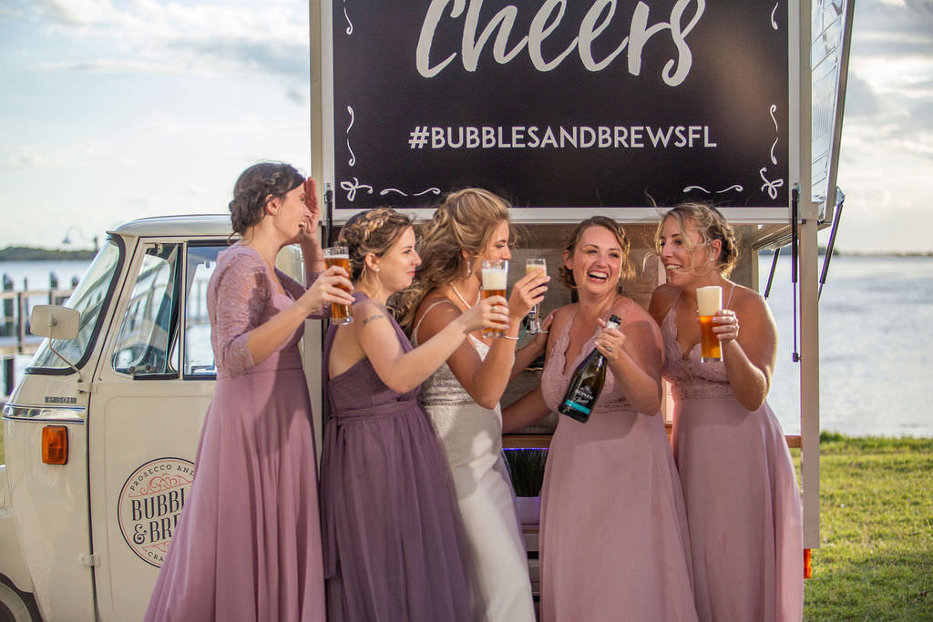 Fun Spring Tampa Bay Bride and Bridesmaids in Mix and Match Purple, Lilac, Lavender Dresses with Drinks from Unique Beverage Cart Get Cozy Bubbles and Brew Cart Waterfront Wedding Portrait