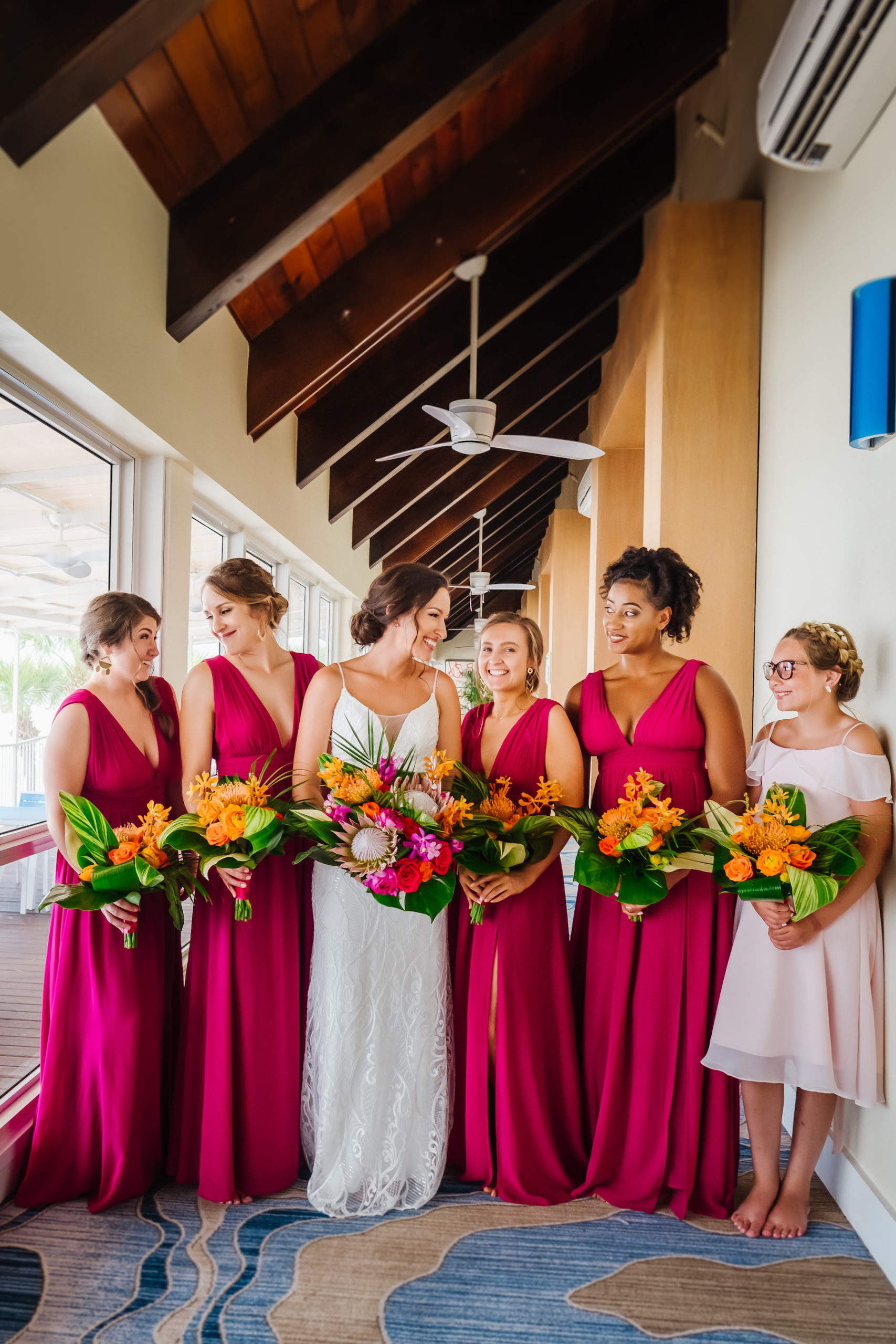 Florida Bride with Bridesmaids in Matching Magenta Dresses Holding Tropical Monstera Palm Leaves, Orange Flowers, King Proteas, Pink and Orange Roses Floral Bouquets | Tampa Bay Waterfront Beach Hotel Wedding Venue Hilton Clearwater Beach