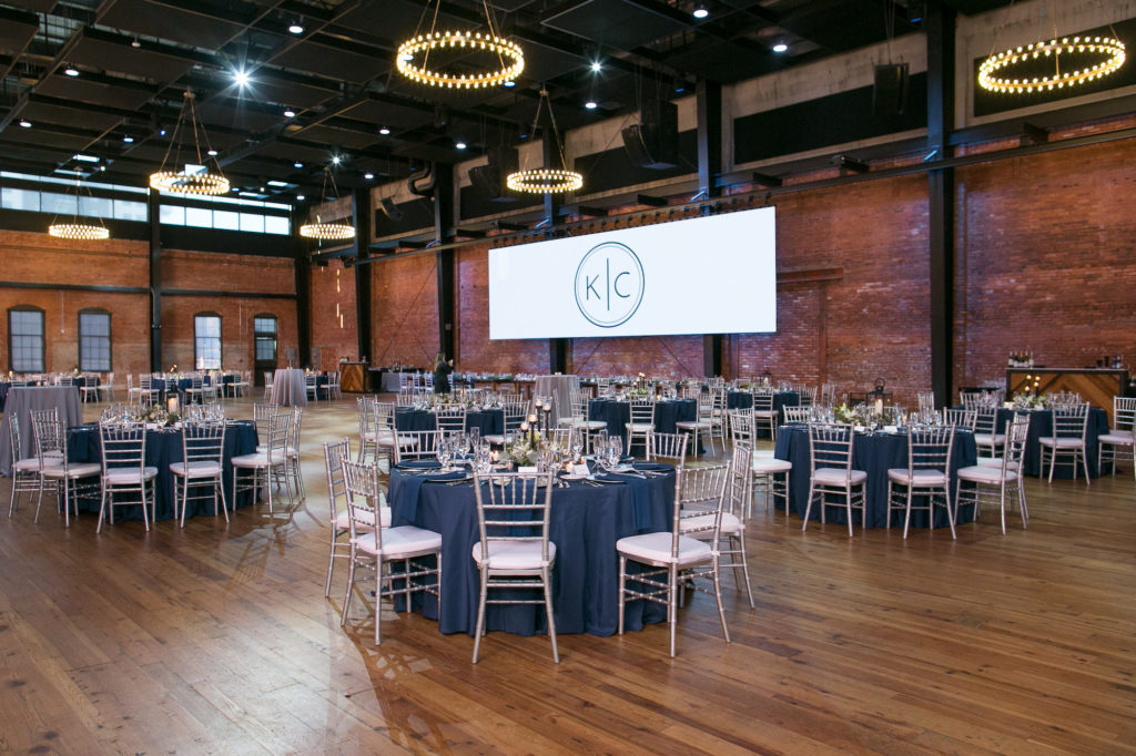 Modern Wedding Reception Decor, Round Tables with Navy Blue Linens, Silver Chiavari Chairs, Round Chandeliers, Custom Monogram Projection | Tampa Bay Wedding Photographer Carrie Wildes Photography | Wedding Planner Love Lee Lane | Historic Industrial Red Brick Wedding Reception Venue Armature Works | Chair and Table Rentals A Chair Affair