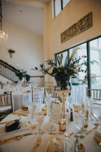 Elegant, Garden Inspired Wedding Decor and Reception, Round Tables with White Linens, Gold Chiavari Chairs, Tall Floral Centerpieces with Green Palms, White Flowers, and Gold Candle Votives, in Clearwater Modern Ballroom with natural lighting, floor to ceiling windows, tall vaulted ceilings, and iron rod staircase | Tampa Bay Wedding Florist Brides N Blooms | Florida Wedding Venue Beso Del Sol | Tampa Wedding Decor Rental Outside the Box Event Rentals