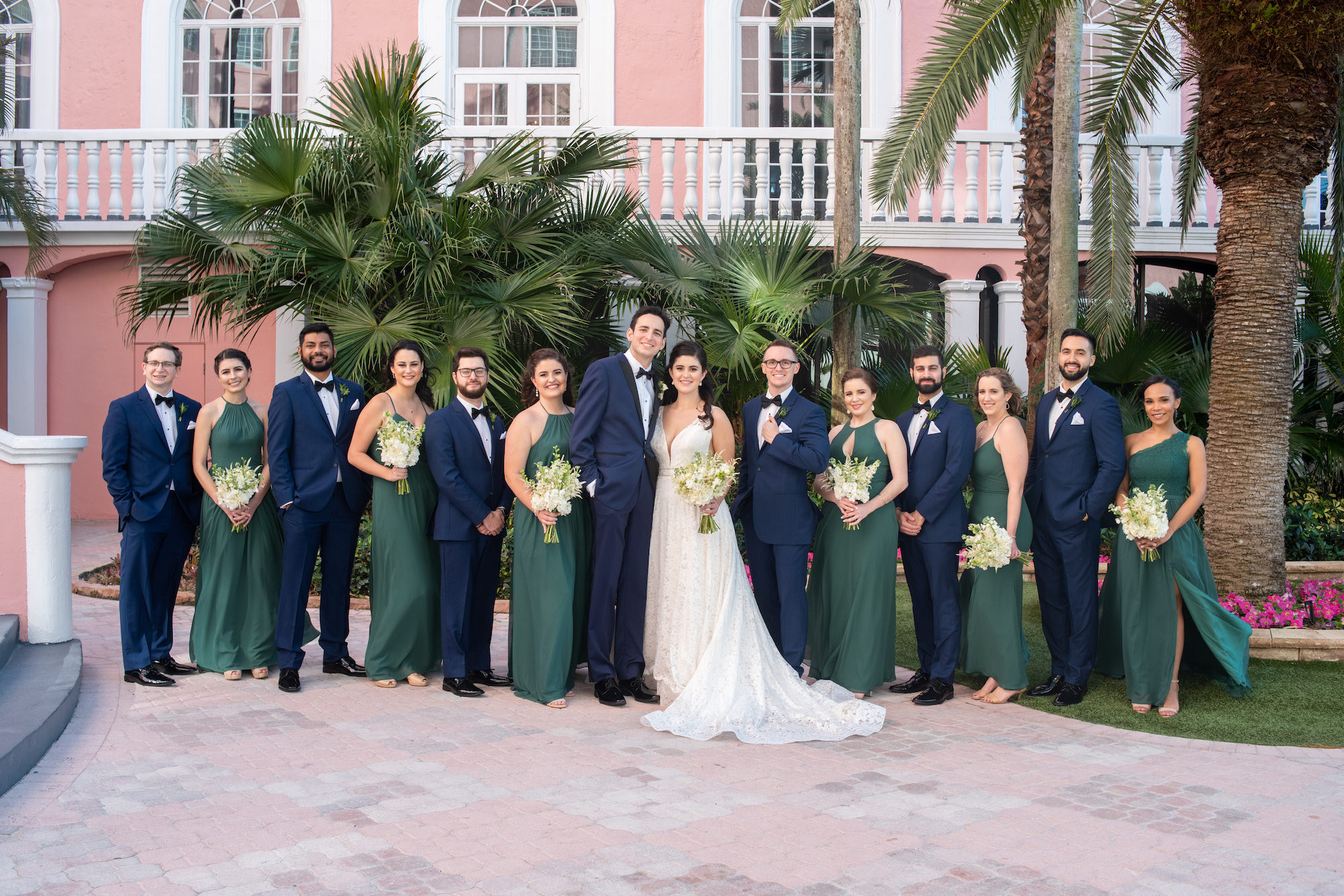 Classic Florida Bride in Lace Paloma Blanca Wedding Dress, Groom and Groomsmen in Navy Blue Suits with Bowties, Bridesmaids in Dark Green Azazie Mix and Match Dresses Holding Ivory Floral Bouquets Wedding Party Portrait | St. Petersburg Waterfront Wedding Venue The Don CeSar | Historic Pink Palace | Tampa Bay Wedding Planner UNIQUE Weddings + Events