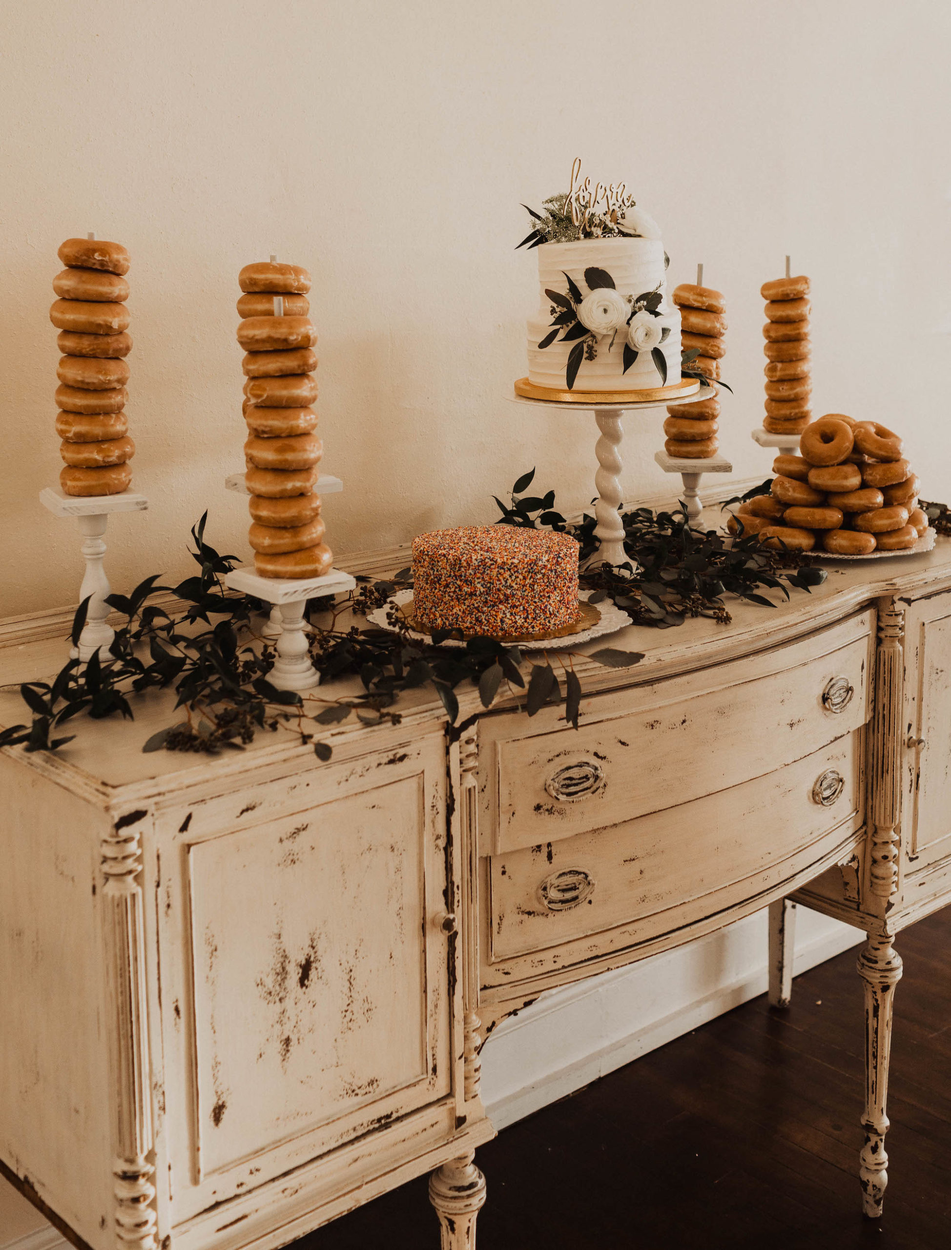 Antique White Dresser Dessert Table Setup, Krispy Kreme Donuts on Spindles, Two Tier White Ruffled Wedding Cake with Roses and Eucalyptus