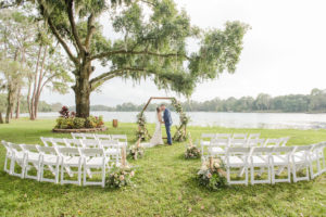 Florida Bride and Groom Kiss During Outdoor Waterfront Wedding Ceremony, Under Large Flowing Oak Tree, Rustic Wooden Hexagon Wedding Arch with Whimsical Inspired White and Dusty Rose and Pompous Grass Ceremony Flowers, White Chairs for Intimate Lakefront Ceremony | Tampa Bay Wedding Planner and Day of Coordinator Elegant Affairs by Design | Venue Barn at Crescent Lake