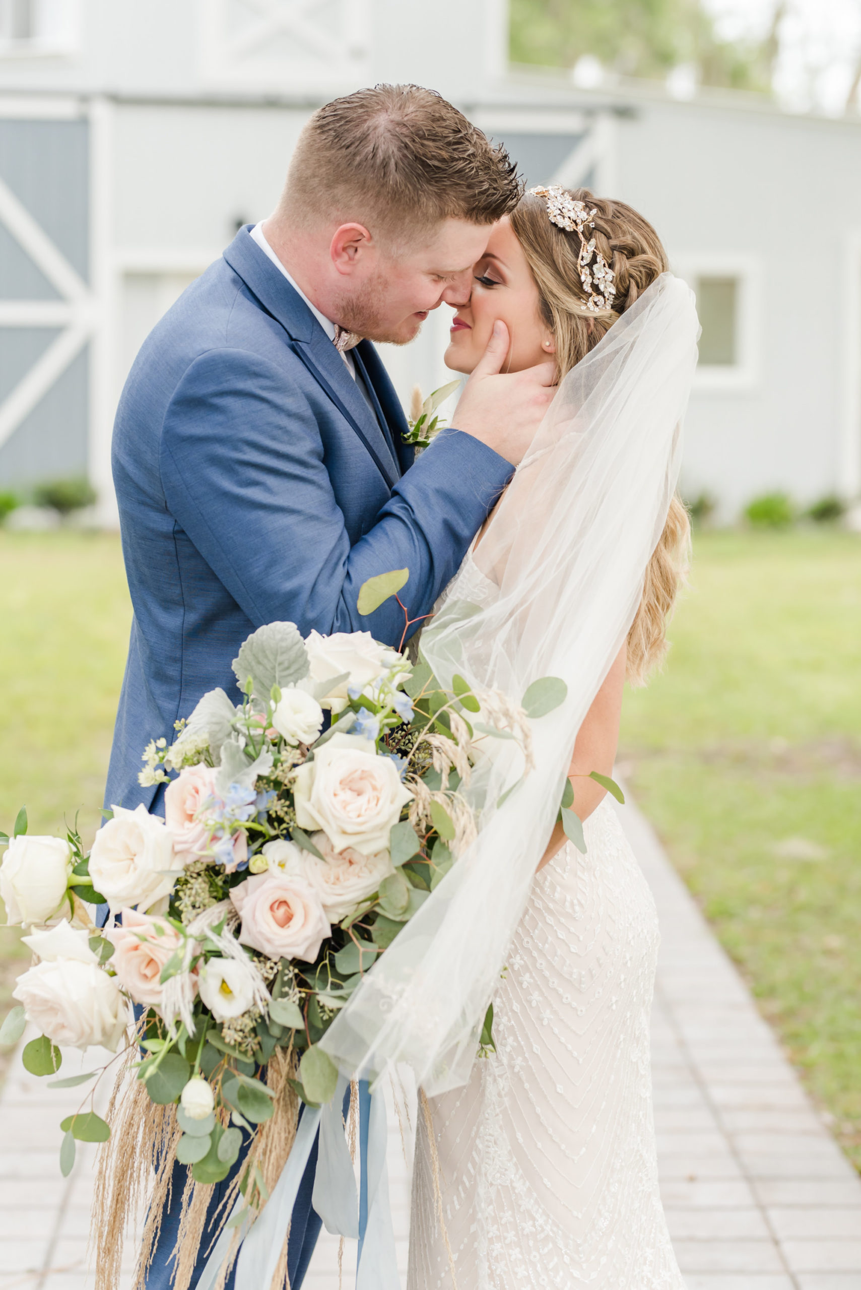 Tampa Bay Bride and Groom Intimate Embrace outside Barn at Crescent Lake, Groom in Dark Blue Suite, Bride holding Bouquet Whimsical Dusty Rose Florals, Ivory Roses, Light Blue Flowers, Pompous Grass, with Eucalyptus Leaves Greenery