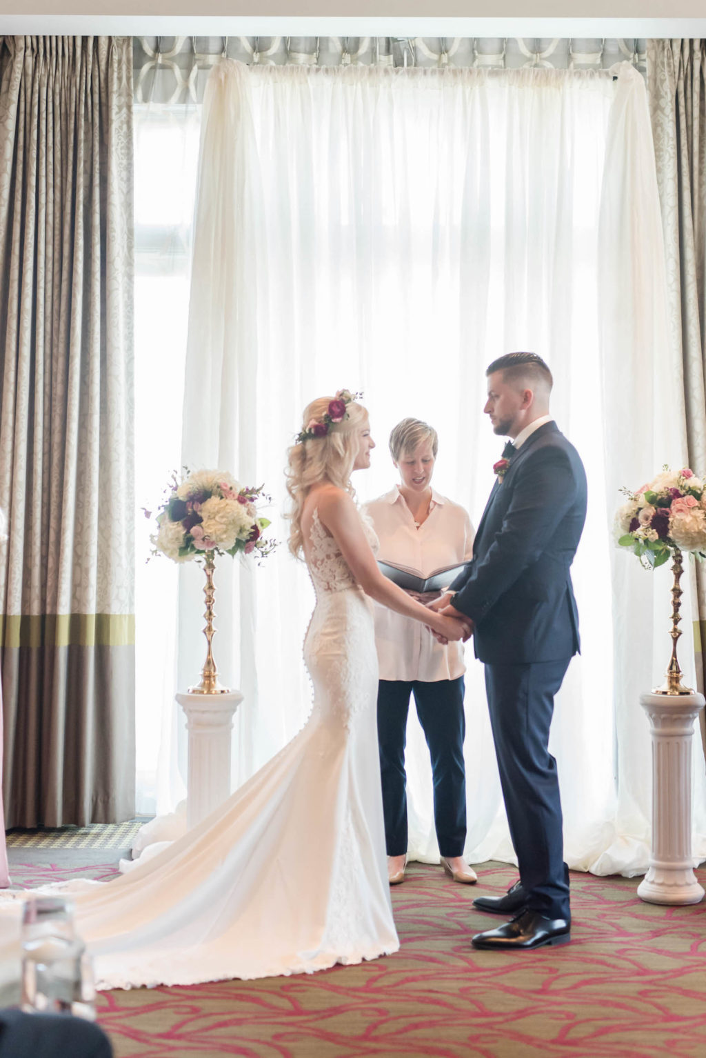 Bride and Groom Exchanging Vows | St. Pete Wedding Venue The Birchwood | Indoor Ballroom Wedding Ceremony with Floral Candlestick and Pipe and Drape Backdrop | Stella York Lace Sheath Spaghetti Strap Empire Waist Wedding Dress Bridal Gown | Groom Classic Black Suit Tux