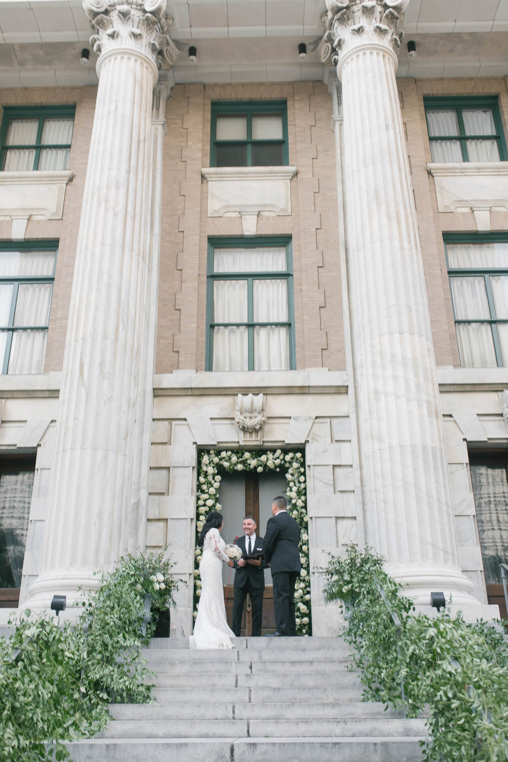 Florida Bride and Groom Exchanging Vows on Steps of Tampa Historic Courthouse Boutique Hotel Wedding Venue Le Meridien, Greenery Decor on Steps, White Roses Doorway Arch | Wedding Planner Parties A'la Carte