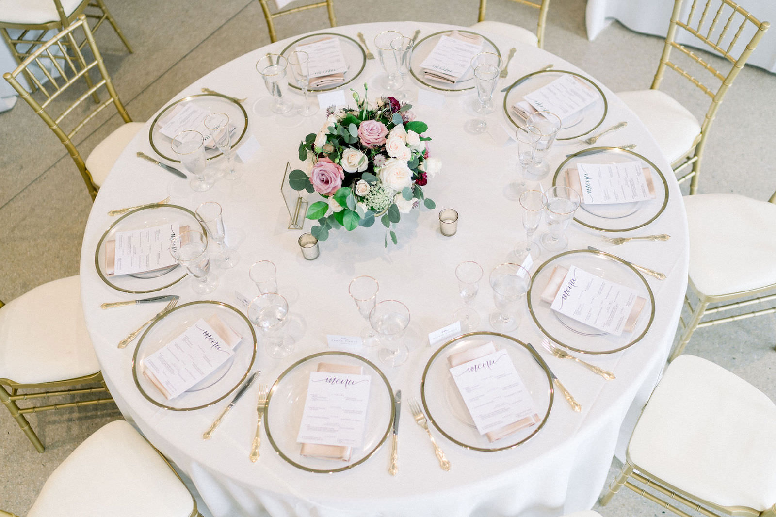 Wedding Reception Table with White Linen Tablecloth and Clear Glass Charger Plates with Gold Rim and Gold Rimmed Glasses and Gold Chiavari Chairs | White Blush Pink and Maroon Red Roses Centerpiece with Eucalyptus Greenery
