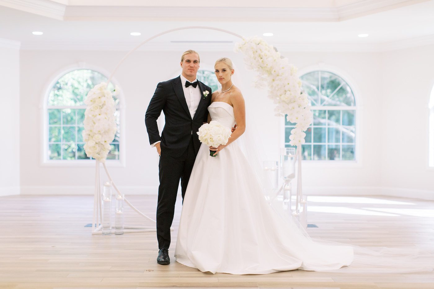 Classic Traditional Bride in Strapless Crepe Romona Keveza Ballgown Wedding Dress Holding White Floral Bouquet, Groom in Black Tuxedo Portrait in Front of Circular Arch with White Orchid Arrangements | Tampa Bay Wedding Planner Parties A'la Carte | Safety Harbor Church Wedding Venue Harborside Chapel