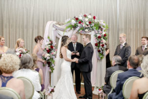 Tampa Bay Bride and Groom Exchange Vows in Ballroom Ceremony, Under Romantic Floral Wedding Arch with Red, White and Mauve Roses, Purple Flowers with Greenery, Ivory Draping | Downtown St. Pete Wedding Planner Blue Skies Weddings and Events | Florida Wedding Photographer Lifelong Photography Studio